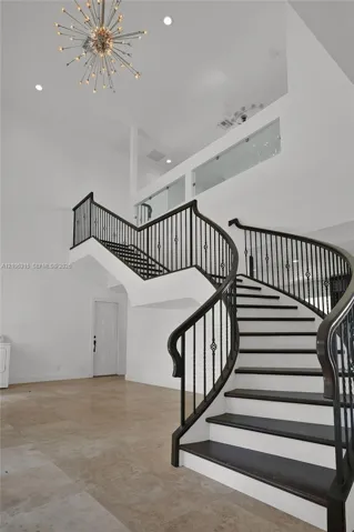 Curved staircase with dark-finished wood treads and metal balusters, polished tile flooring, double-height ceiling, glass-panel balcony railing, and starburst-style chandelier