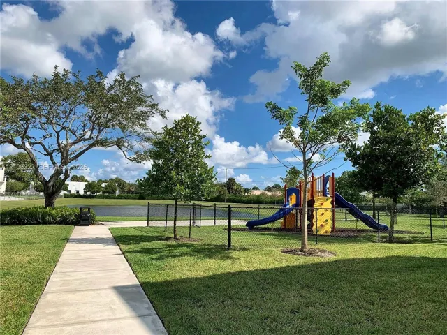Community grounds featuring a concrete pathway, a playground structure, mature trees, open green space, and a pond