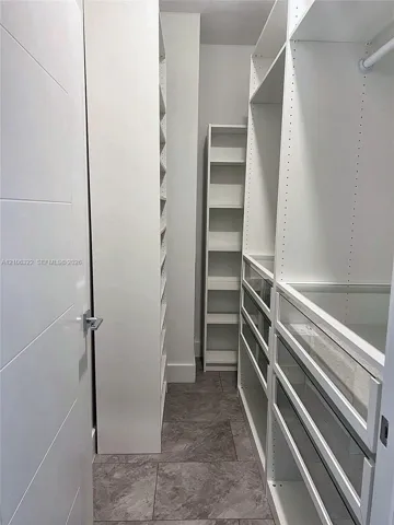 Custom walk-in closet featuring white built-in shelving, glass-front drawers, hanging rods, and stone-finish tile flooring
