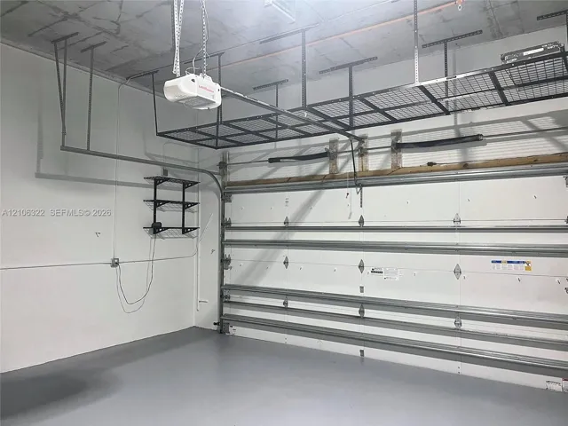Garage space featuring finished epoxy flooring, white paneled walls, exposed concrete ceiling, integrated overhead metal storage racks, and wall-mounted metal shelving