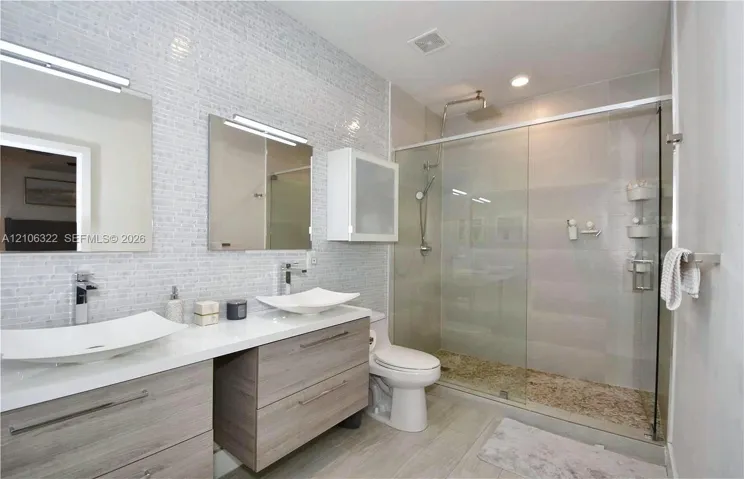 Dual floating vanity with rectangular vessel sinks, chrome faucets, wall-mounted medicine cabinet, and a walk-in glass-enclosed shower featuring a textured stone-finish floor