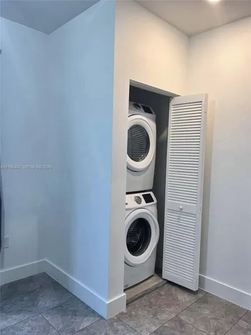 Laundry closet featuring stacked appliances and a white louvered door, complemented by grey tile flooring