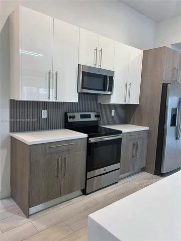 Kitchen featuring dual-tone cabinetry with gloss-white uppers and textured wood-finish lowers, white solid-surface countertops, vertical-slat backsplash, stainless steel appliances, and plank-style flooring
