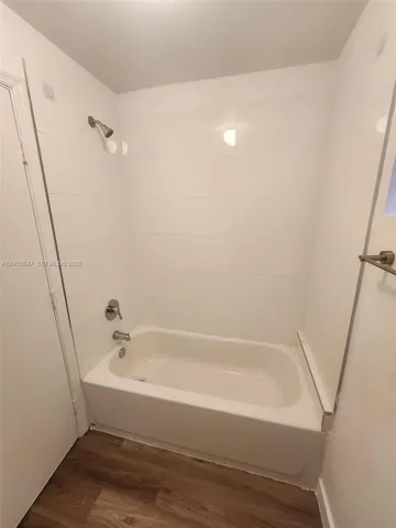 Bathtub and shower combination featuring large-format white tile surround, metallic plumbing fixtures, and wood-finish flooring