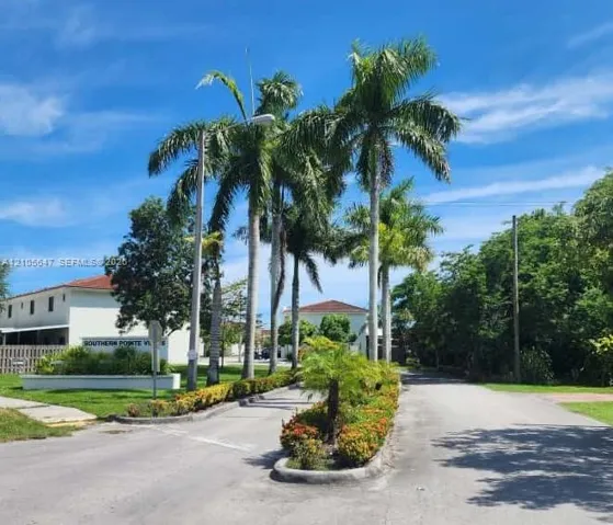 Entryway featuring paved roads, mature palm trees, manicured landscaping, and building exterior with a tile roof