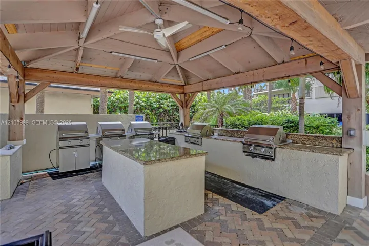 Covered outdoor kitchen featuring a timber-framed vaulted ceiling, stone countertops, multiple built-in stainless steel grills, and herringbone brick paver flooring
