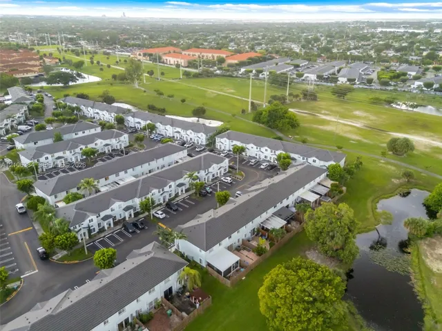 Aerial perspective of residential community featuring multi-unit structures, private parking areas, landscaped grounds, mature trees, and a water feature