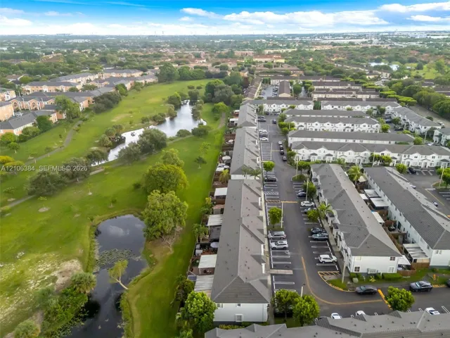 Aerial perspective of a multi-unit residential community featuring pitched shingle roofs, paved asphalt parking areas, expansive common green space, and natural water features