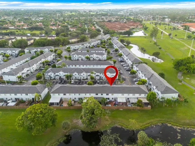 Aerial perspective of multi-family residential architecture featuring pitched shingled roofing, light-toned siding, and paved parking surfaces, situated within a landscape of expansive green space and water features