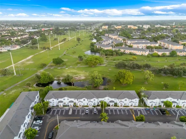 Aerial perspective of multi-family structures featuring peaked grey-tone roofs and white siding, including dedicated asphalt parking areas and proximity to expansive green space with mature trees and a retention pond