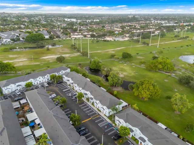 Aerial perspective of a multi-unit residential development featuring a gray shingle roofline, paved parking area with designated stalls, and extensive green space with established trees and water features
