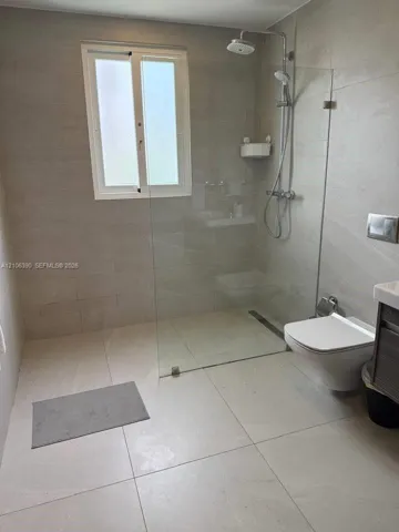 Walk-in shower with a glass panel, rainfall shower head, wall-mounted toilet, and large-format tile flooring and wall surfaces