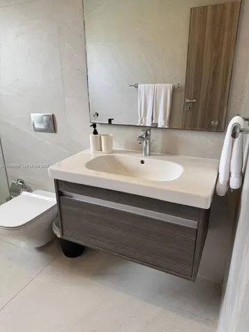 Floating wood-finish vanity with an integrated basin, wall-mounted flush toilet, large frameless mirror, and large-format tile flooring