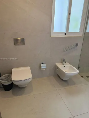 Wall-mounted white toilet, wall-mounted white bidet with chrome faucet, large-format stone tile flooring and wall cladding, and chrome wall-mounted flush actuator