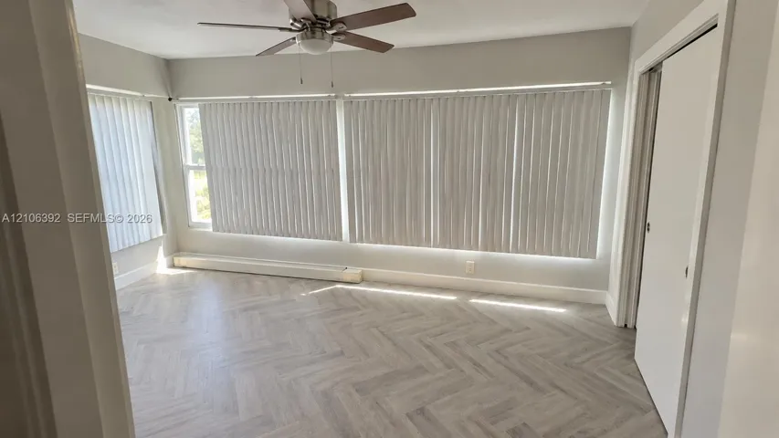 Herringbone-pattern wood-finish flooring, extensive floor-to-ceiling windows with vertical blinds, and a neutral-toned ceiling fan with integrated lighting