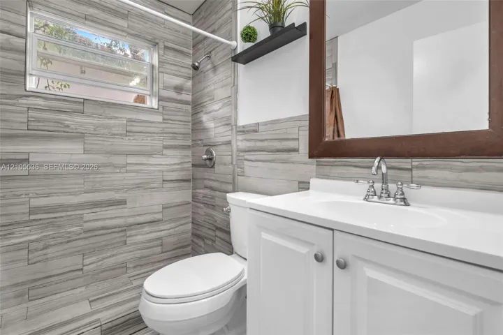 Bathroom featuring horizontally-laid tile wall treatments, white cabinetry with an integrated basin, a polished chrome double-handle faucet, and an awning-style window