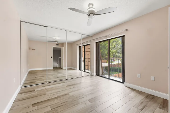 Wood-finish flooring, full-wall mirrored closet doors, ceiling fan with integrated lighting, and sliding glass doors opening to an outdoor balcony