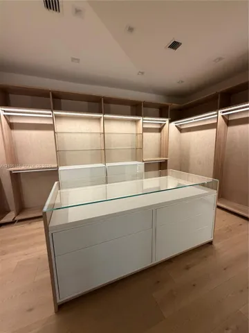 Custom Women’s primary closet , long mirror ,built-in wood cabinetry featuring integrated lighting and glass shelving, centered by a matching island with a glass top and matte finish drawer fronts, complemented by wood-finish flooring and recessed ceiling lighting