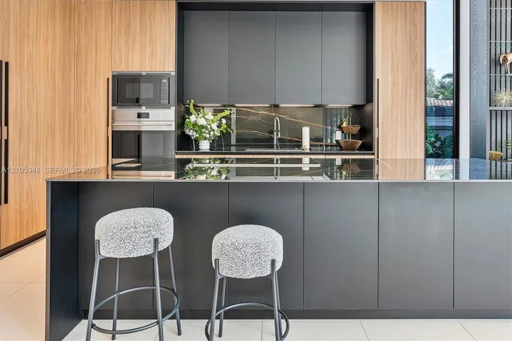 Modern kitchen featuring matte charcoal cabinetry, wood-finish paneling, an expansive island with a stone surface, built-in stainless steel appliances, and matching stone backsplash