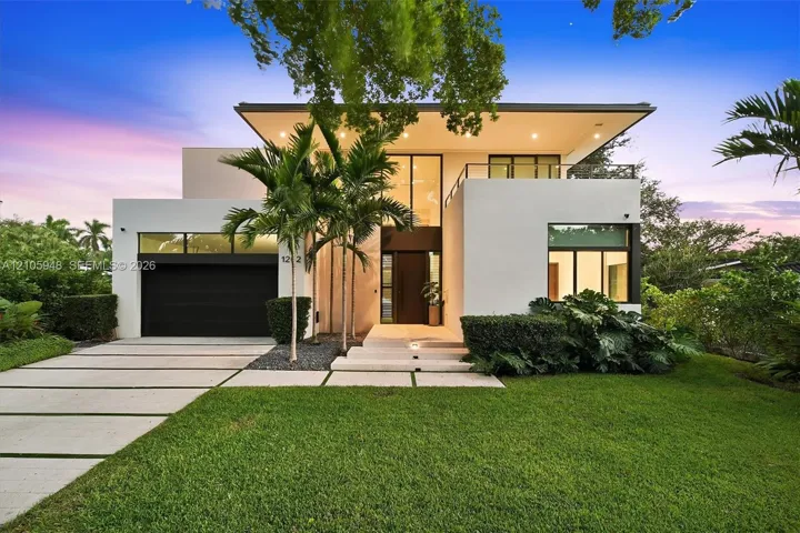 Modern architectural exterior featuring a flat roofline, oversized glass entry, second-level balcony with metal railing, paver driveway, and recessed exterior lighting