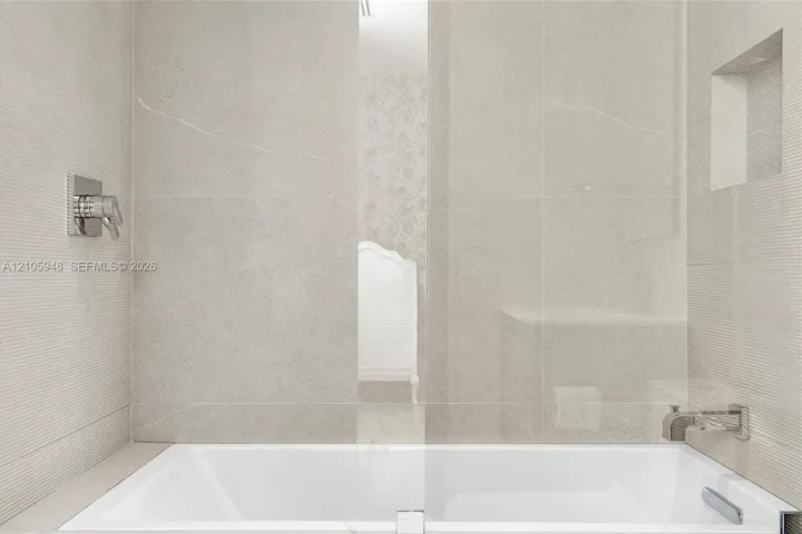 Soaking tub featuring large-format neutral tile surround, integrated wall niche, chrome wall-mounted faucet, and glass partition