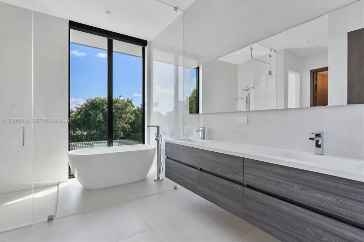 Modern bathroom featuring a freestanding soaking tub, dual-sink wall-mounted vanity with dark wood-finish cabinetry, white solid-surface countertop, floor-to-ceiling glass paneling, and large-format tile flooring