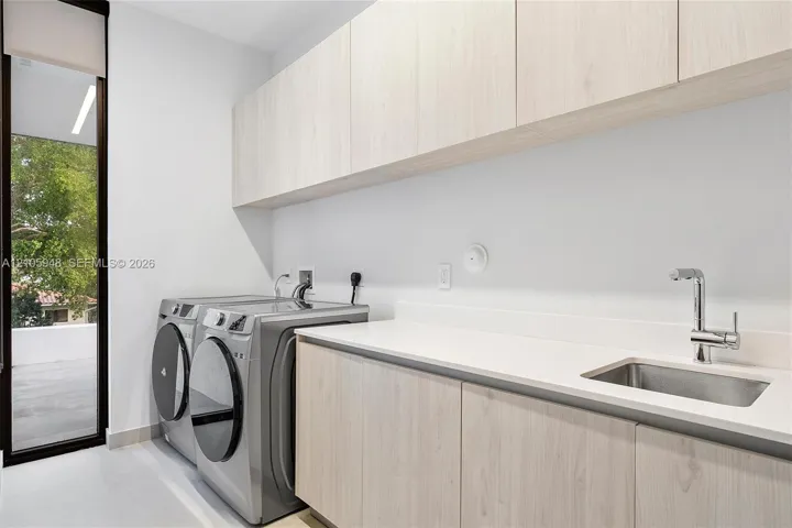 Laundry space featuring light-toned cabinetry, solid-surface countertops, an integrated stainless steel sink with a high-arch faucet, and full-height glass exterior door