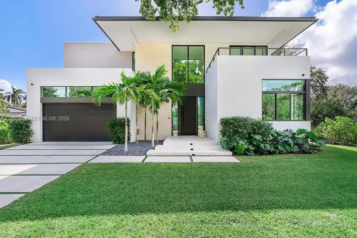 Modern architectural exterior featuring smooth stucco finish, geometric flat roofline, oversized floor-to-ceiling windows, second-story balcony with metal railing, and paver driveway with integrated turf strips