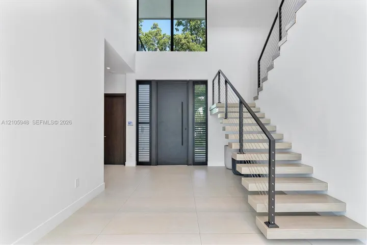 Grand foyer featuring a double-height ceiling, floor-to-ceiling windows, modern floating staircase with cable railing, large-format tile flooring, and a sleek oversized entry door with side lites