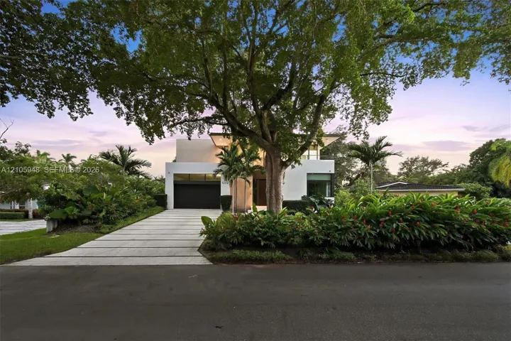 Modern architectural facade featuring a smooth stucco exterior, linear concrete paver driveway, dark-finish multi-car garage door, and lush tropical landscaping with mature canopy trees
