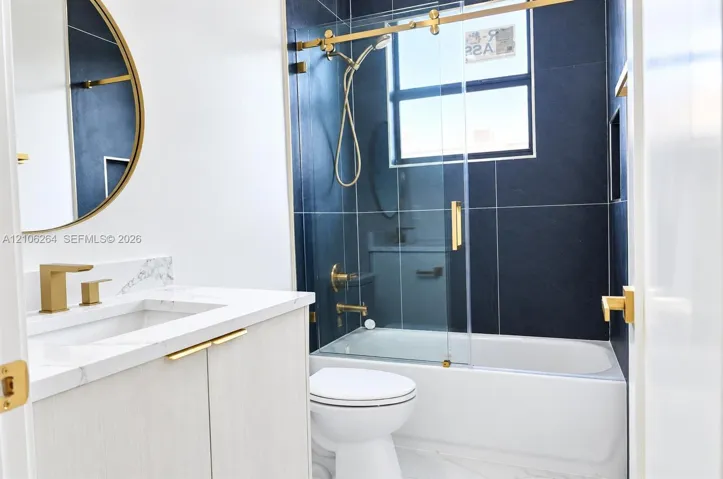 Bathroom featuring a light wood-finish vanity with a stone countertop, a brass-rimmed circular mirror, gold-tone fixtures, and a glass-enclosed shower with dark tile surround