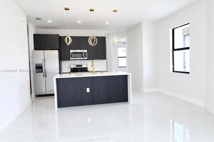 Kitchen featuring a central island with fluted cabinetry and a solid-surface countertop, dark-toned cabinetry, stainless steel appliances, polished tile flooring, and metallic spherical pendant lighting