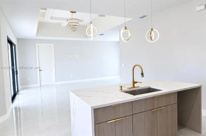 Kitchen island featuring a waterfall-edge countertop, undermount stainless steel sink with gold-tone faucet, wood-finish cabinetry with gold-tone hardware, geometric ring pendant lighting, and tray ceiling detail