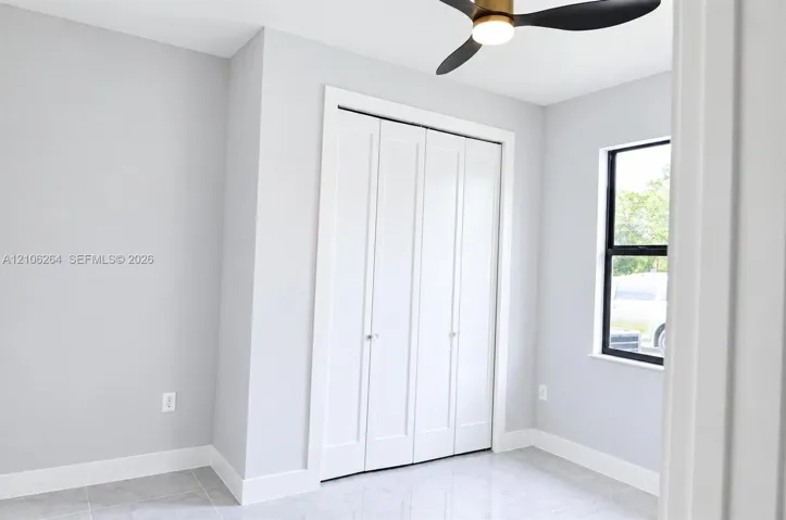 Neutral interior featuring light-toned tile flooring, four-panel folding closet doors, a black-framed window, and a modern brass and black finish ceiling fan with integrated lighting