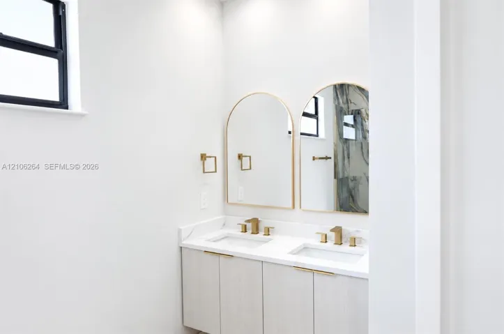 Dual-sink vanity featuring light wood-finish cabinetry, white stone-surfaced countertops, brushed gold-tone faucets, and twin arched mirrors