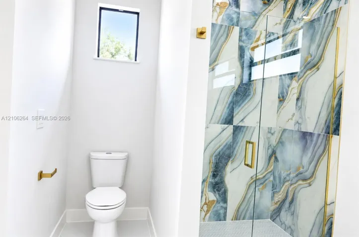 Shower enclosure featuring large-format decorative tile with gold-tone metal trim and hardware, clear glass paneling, and a high-efficiency toilet