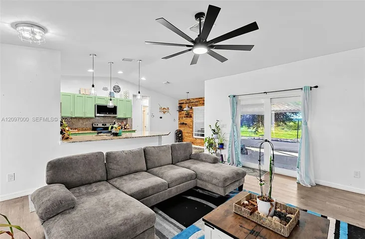 Open-concept interior featuring wood-finish flooring, vaulted ceiling with a multi-blade matte black fan, kitchen breakfast bar with stone-finish countertops, and dual sliding glass doors