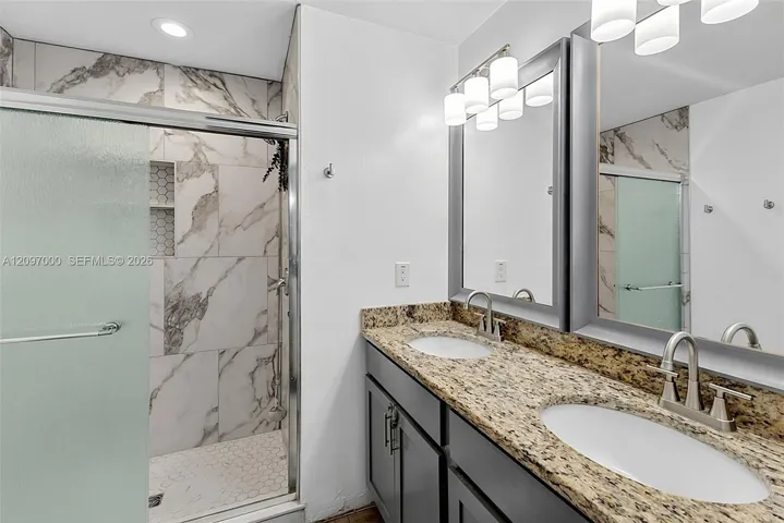 Dual vanity featuring stone-finish countertops, undermount sinks, chrome fixtures, and a walk-in shower with stone-patterned wall tile and recessed shelving