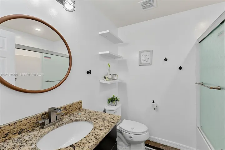 Bathroom featuring a vanity with a stone-finish countertop, undermount sink, round-frame mirror, wall-mounted floating shelving, and a shower enclosure with frosted glass panels