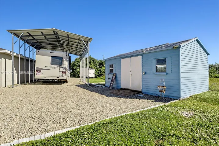 Metal carport structure situated on a gravel surface, complemented by a detached storage building with horizontal siding and double-door entry