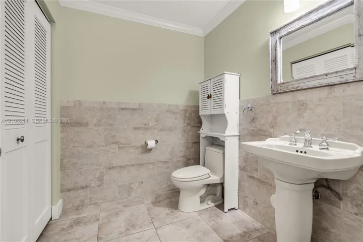 Pedestal sink with polished chrome fixtures, neutral-toned stone-finish tile wainscoting, porcelain tile flooring, crown molding, and white louvered-style closet doors