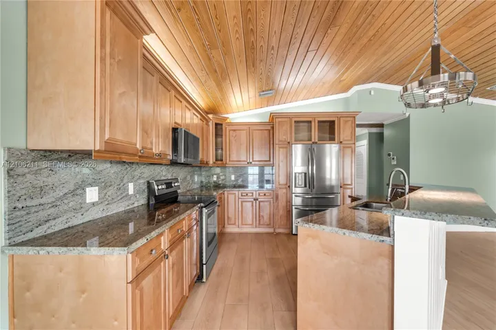 Kitchen featuring natural wood-finish cabinetry, stone slab countertops, stainless steel appliances, vaulted wood-paneled ceiling, and a hanging metal light fixture