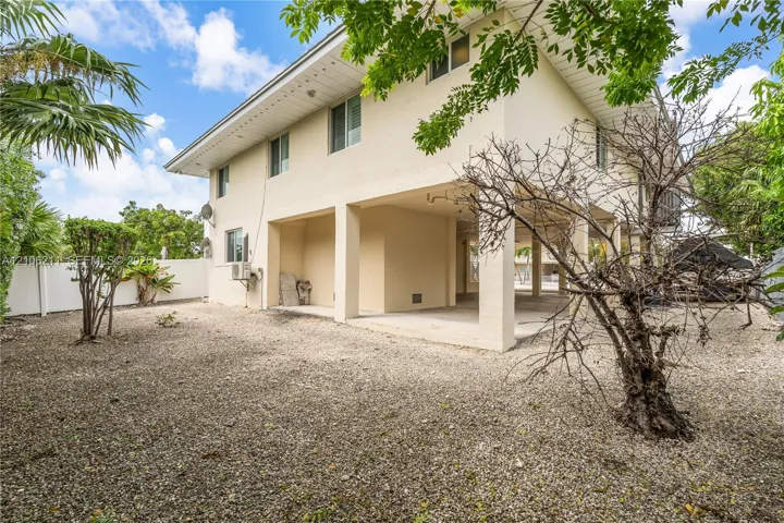 Two-story exterior featuring a cream-colored stucco facade, raised construction with open-air lower level, gravel grounds, and perimeter privacy fencing
