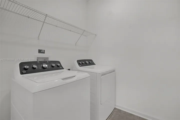 Dedicated laundry area featuring white top-load washer and dryer set, white wall-mounted wire shelving, and neutral tile flooring