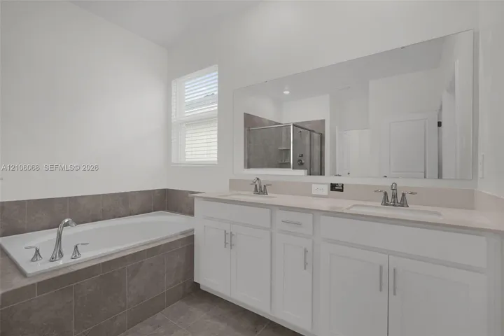 Dual-sink vanity with light-toned countertop, soaking tub featuring tile surround, oversized wall-mounted mirror, and tile flooring