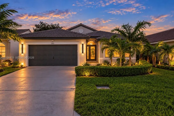 Single-story residence featuring a tile roof, concrete driveway, attached multi-car garage, manicured landscaping, and exterior wall-mounted lighting