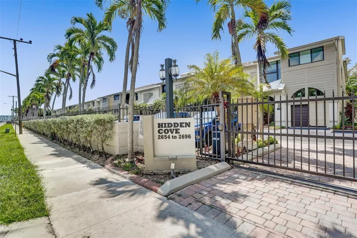 Gated entrance featuring a metal perimeter fence, paver driveway, signage, and mature palm trees