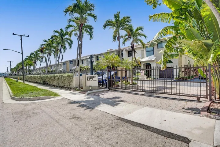 Gated community entrance featuring paver-stone driveway, perimeter metal fencing, and mature palm landscaping