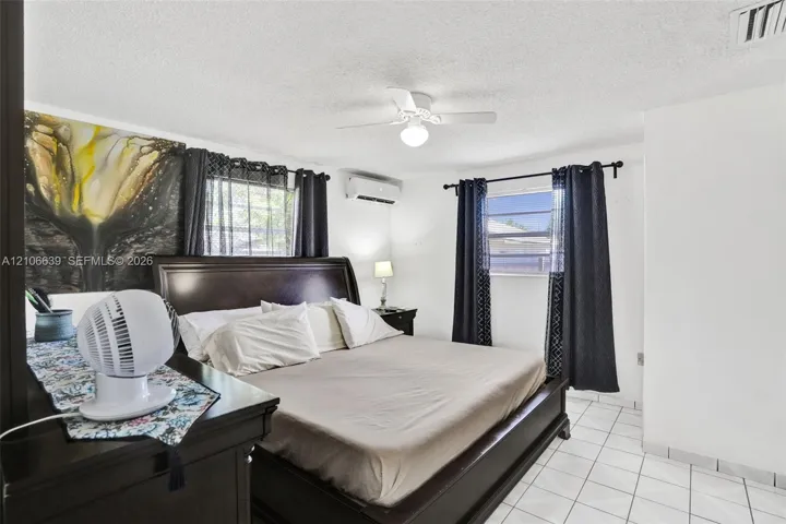 Bedroom featuring white tile flooring, ceiling fan with integrated light fixture, wall-mounted air conditioning unit, and dual windows