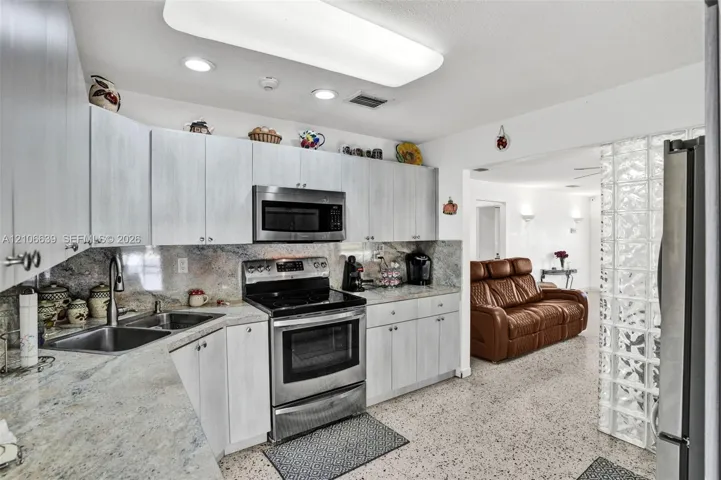 Kitchen featuring light-finish cabinetry, stone countertops, stainless steel appliances, a double-basin stainless steel sink, and a decorative glass block partition